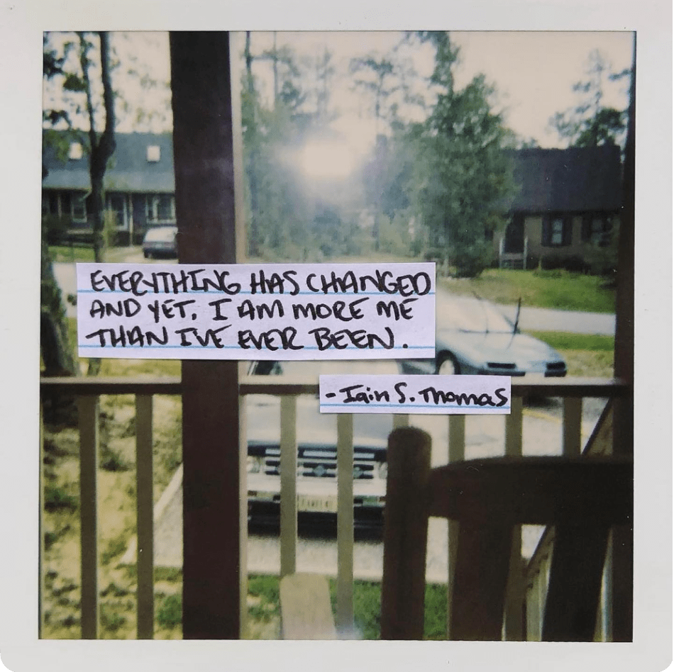 A faded photograph of two cars parked in a driveway, taken from a balcony. Writing overlaid reads: "EVERYTHING HAS CHANGED AND YET, I AM MORE ME THAN I'VE EVER BEEN. –Iain S. Thomas"