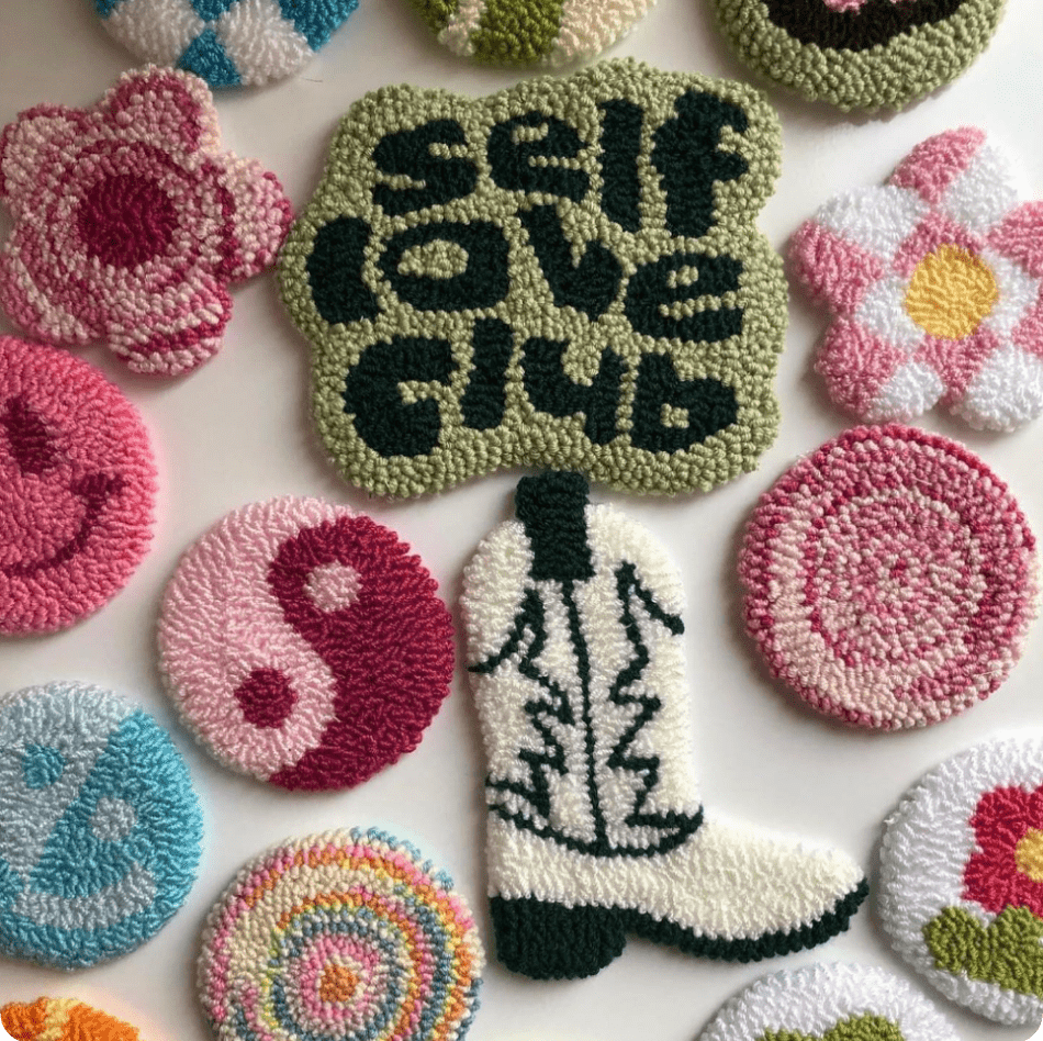 A collection of tiny, colorful patches made of a rug-like material: flowers, smiley faces, a yin-yang symbol, and a cowboy boot. In the center, one of the patches reads: "self love club".