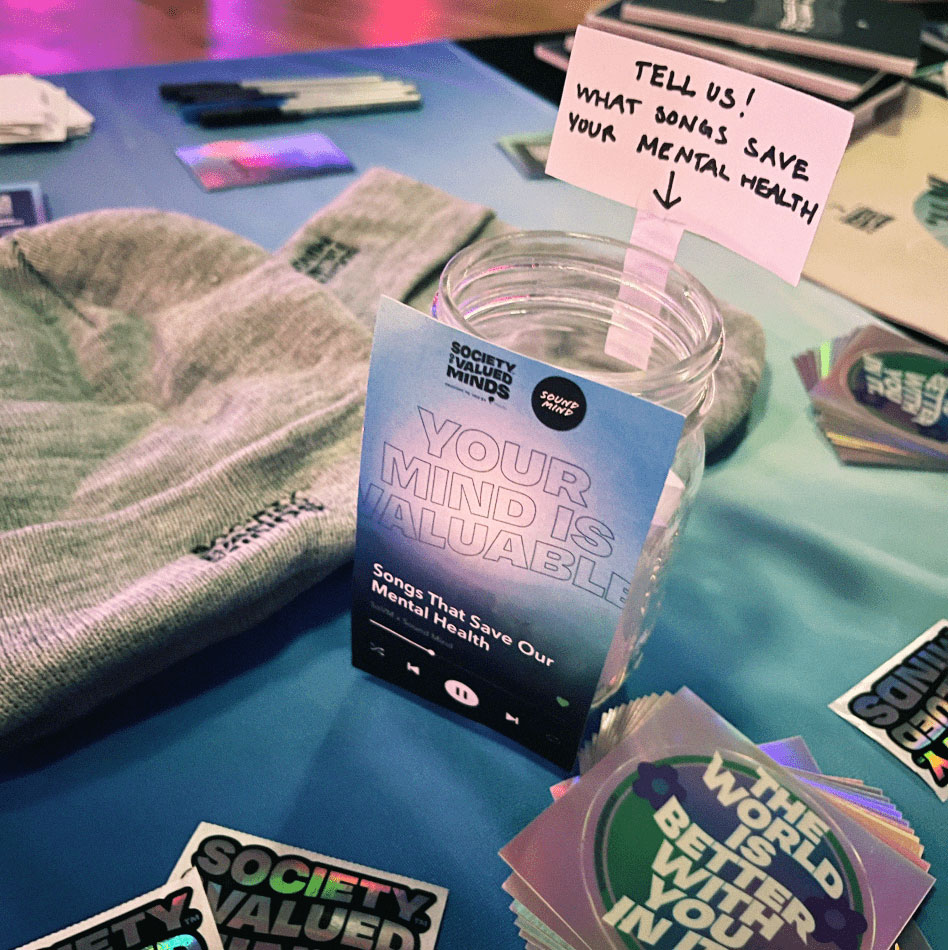 A jar sits on a Society of Valued Minds table at a Sound Mind event. An instruction above the jar reads: "TELL US! WHAT SONGS SAVE YOUR MENTAL HEALTH".