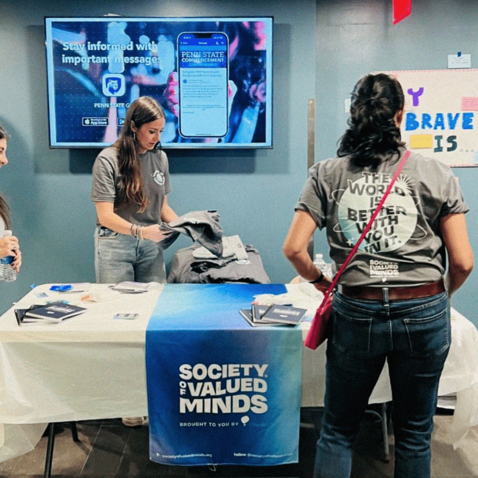A table at an event with a Society of Valued Minds banner draped over it. Stickers, print resources, and journals sit on the table. Someone stands in front of the table with their back to the camera. The back of their shirt reads, "THE WORLD IS BETTER WITH YOU IN IT."
